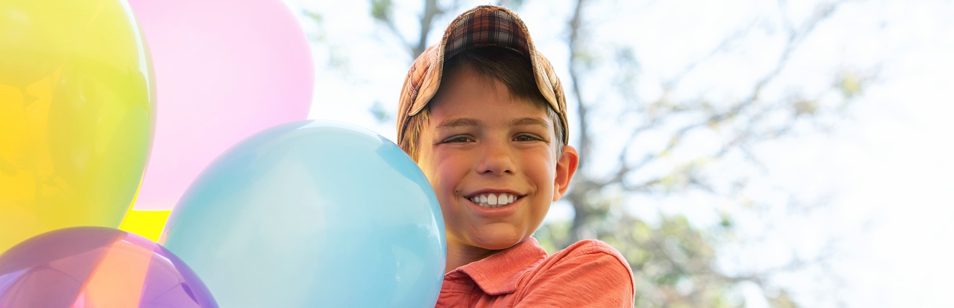 Ein breit grinsender Junge in einem karrierten Cap steht neben ein paar in der Luft stehenden Luftballons.